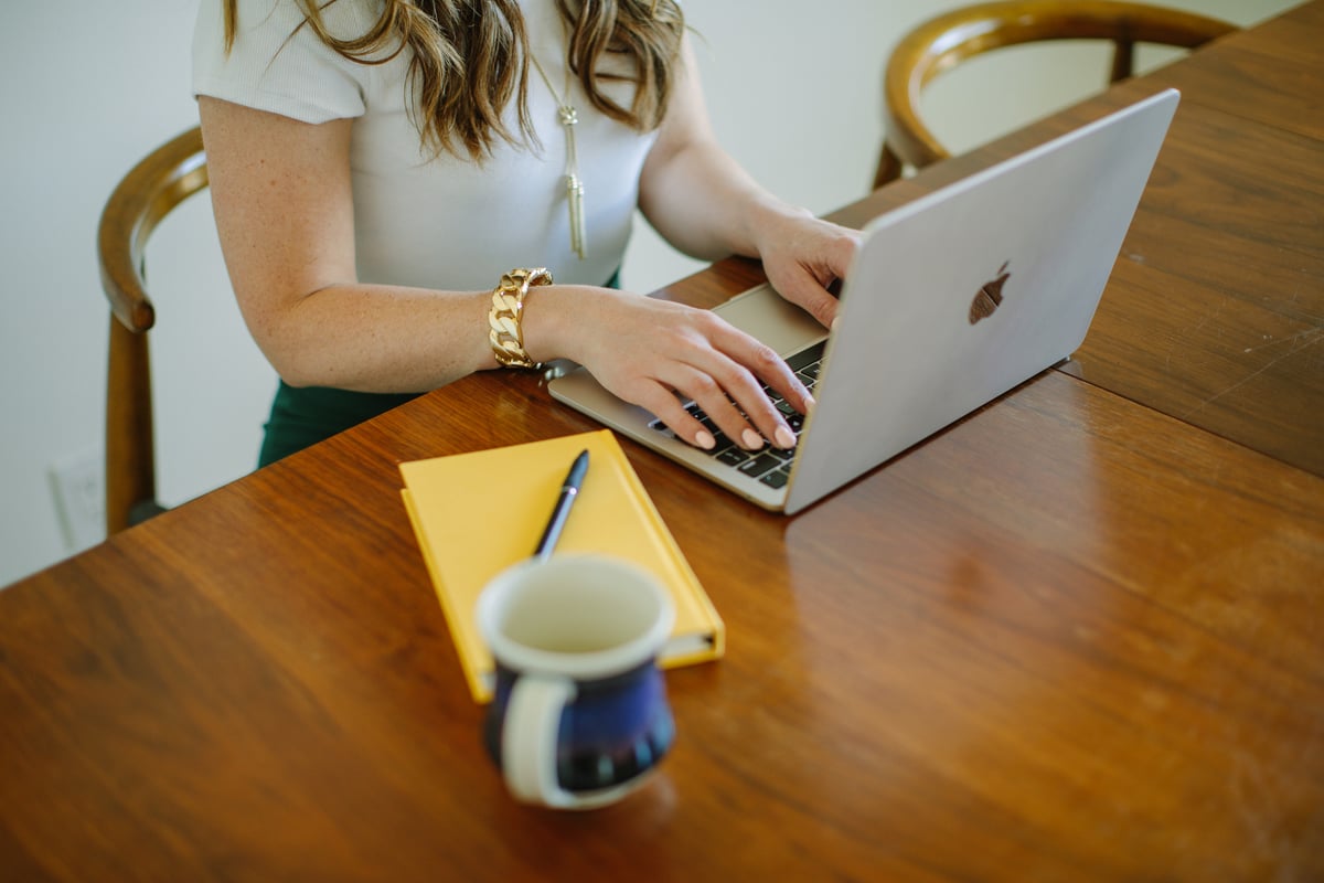 A laptop open on a wooden desk with a yellow journal and coffee mug alongside — telehealth therapy at home.